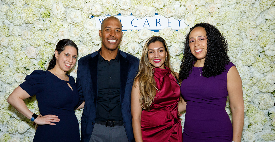 Three women and one man, all W. P. Carey employees, smiling in front of a wall covered in white roses and a small white and blue sign saying "W. P. Carey".