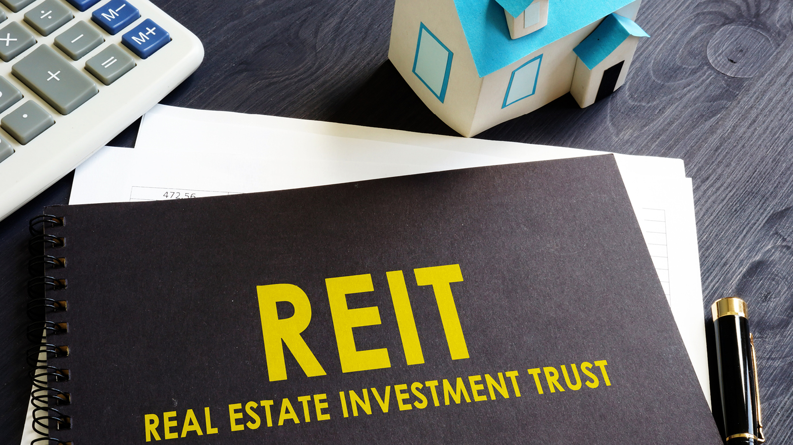 A black folder labeled REIT (Real Estate Investment Trust) in yellow type on a desk with a calculator and a toy house