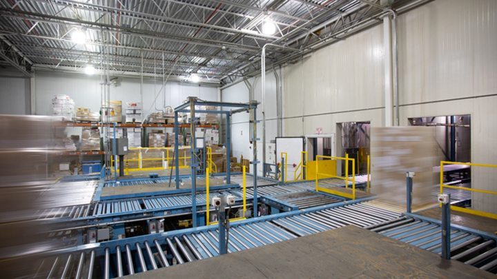 The interior of a warehouse with a metal roof, blue machines and cardboard boxes moving quickly along the machines.