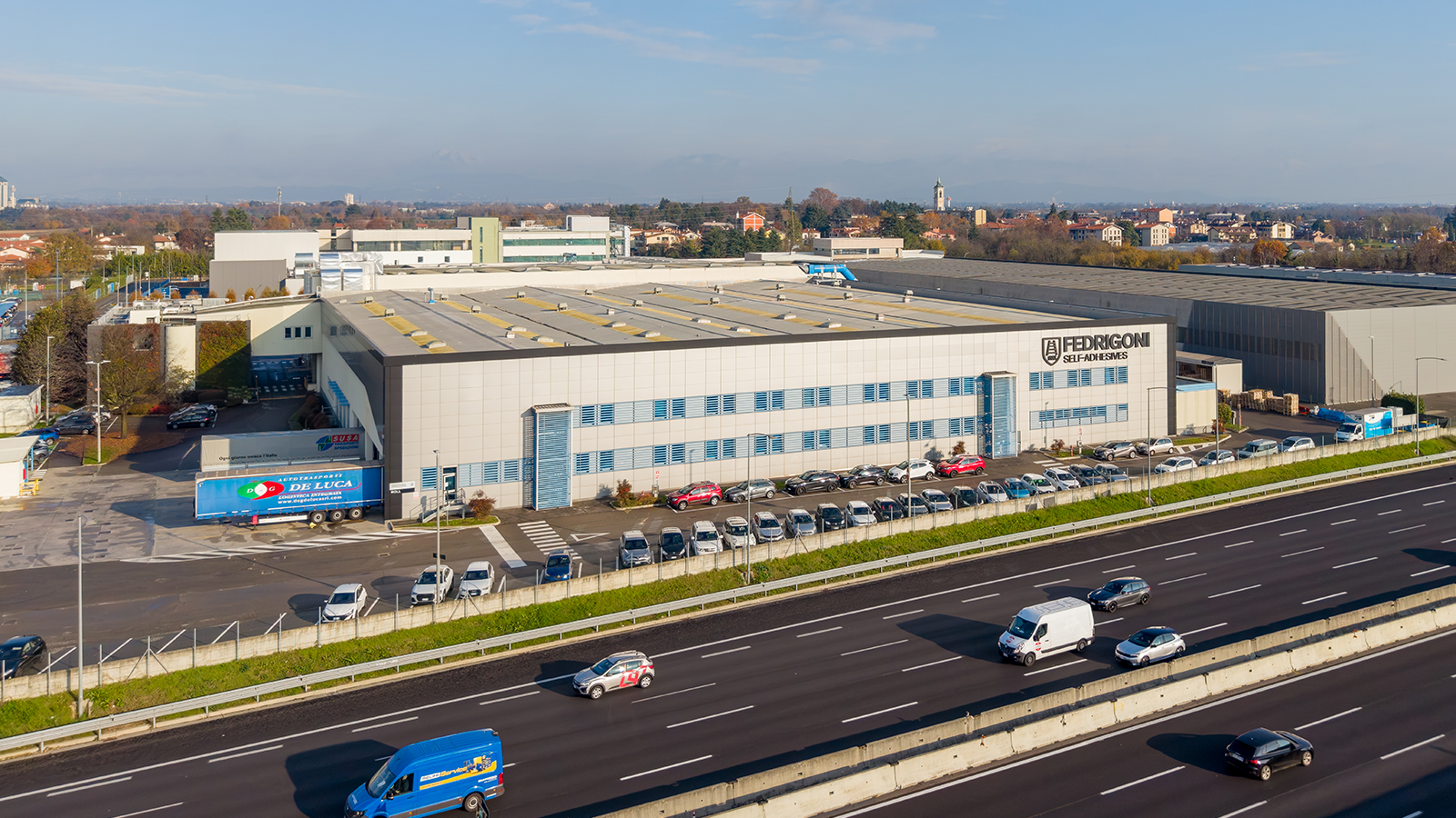 A warehouse leased to Fedrigoni that sits in front of a highway, with a packed parking lot and a blue sky