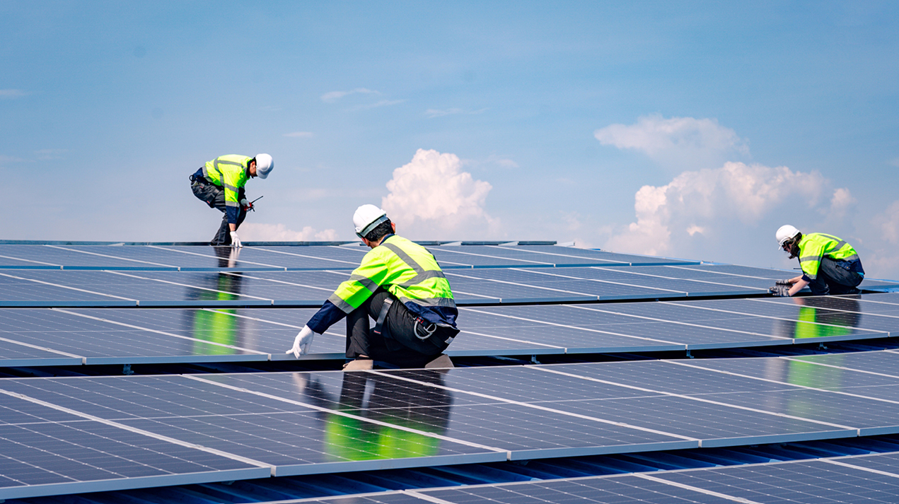 Three men wearing reflective gear on the top of a row of solar panels with a clear sky behind them.