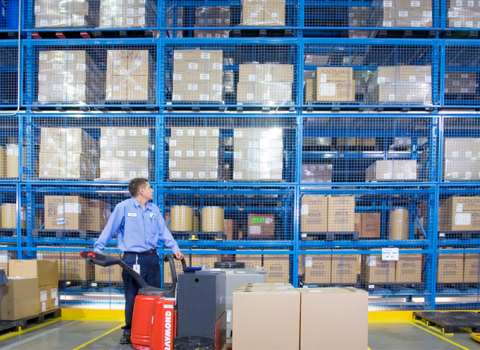 Inside of a W. P. Carey (WPC) net-leased warehouse with crowded blue shelves and a worker wearing a blue shirt