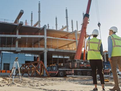A photo of 3 people with their backs to the camera wearing hard hats and hi-vis vests, standing in front of a building under construction