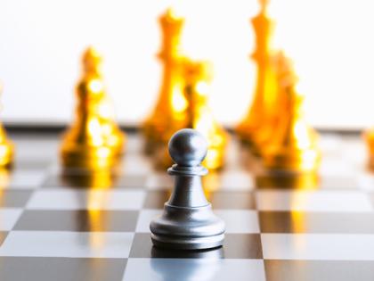 A black pawn sits on a chess board facing a number of opposing gold chess pieces