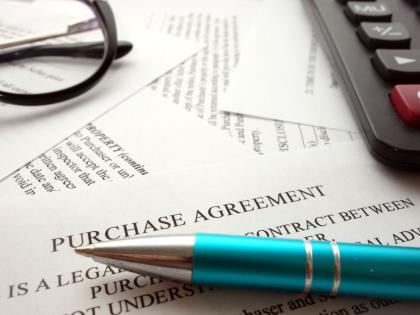 Glasses, a calculator, and a blue pen lying atop a messy pile of papers with the foremost saying "Purchase Agreement" across the top.