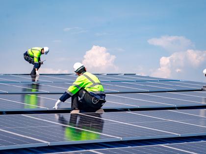 Three men wearing reflective gear on the top of a row of solar panels with a clear sky behind them.