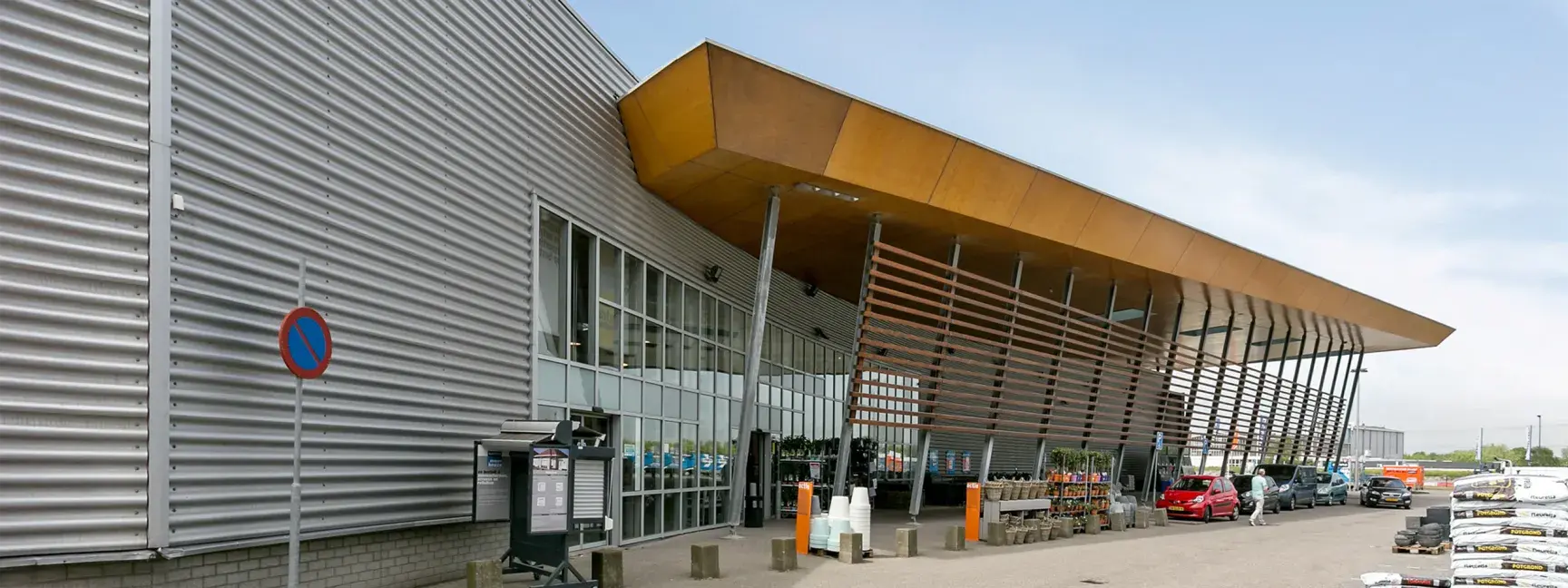 Exterior of a building, metal siding with a wooden roof outcropping covering the sidewalk. Plants are for sale on the sidewalk.
